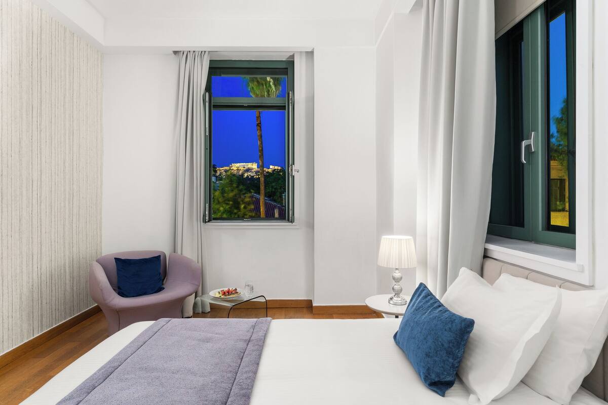 A hotel room with a bed, a chair, a small table with a lamp, and a window with curtains.
