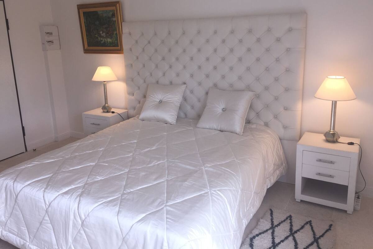 A neatly made bed with a quilted white duvet, two bedside tables with lamps, a white headboard, and a framed picture on the wall.