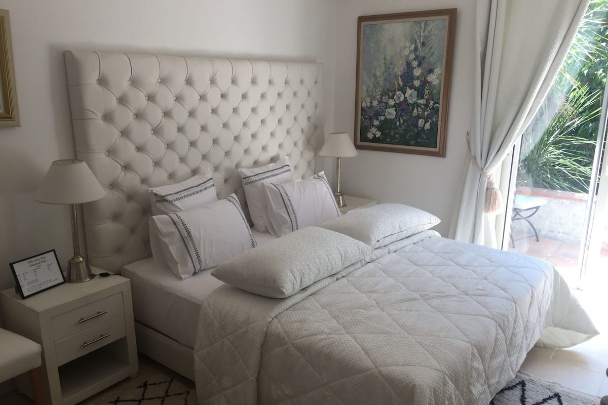 A bedroom with a tufted headboard, white bedding, a bedside table with a lamp, and a framed painting on the wall.