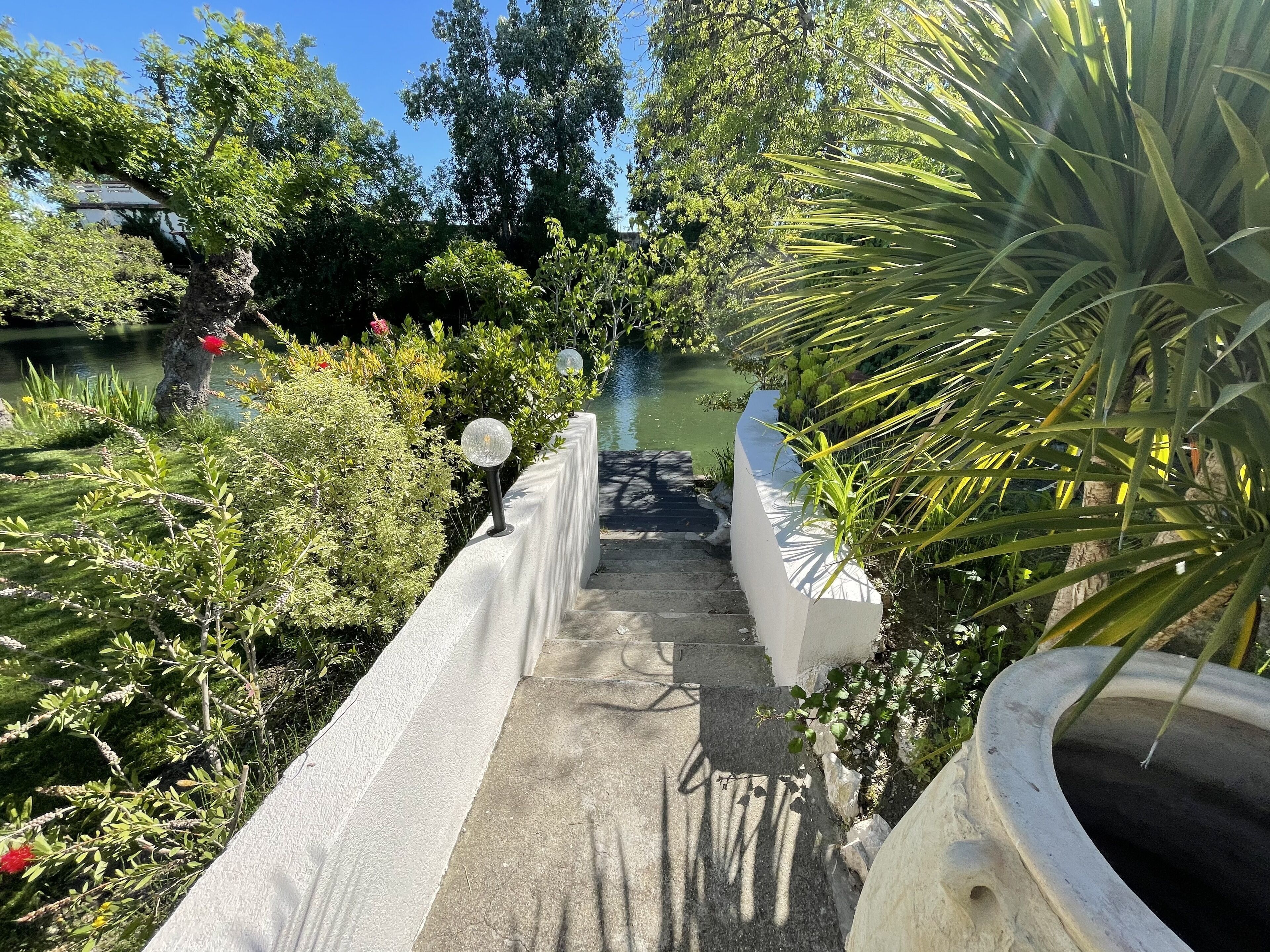 Un sentier qui mène à un étang bordé d’une végétation luxuriante et entouré d’un mur blanc.