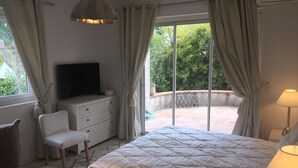 A bedroom with a bed, a TV on a dresser, a chair, and a view of the outdoors through large windows. - Villa Louise-Rose (Villeneuve-Loubet)