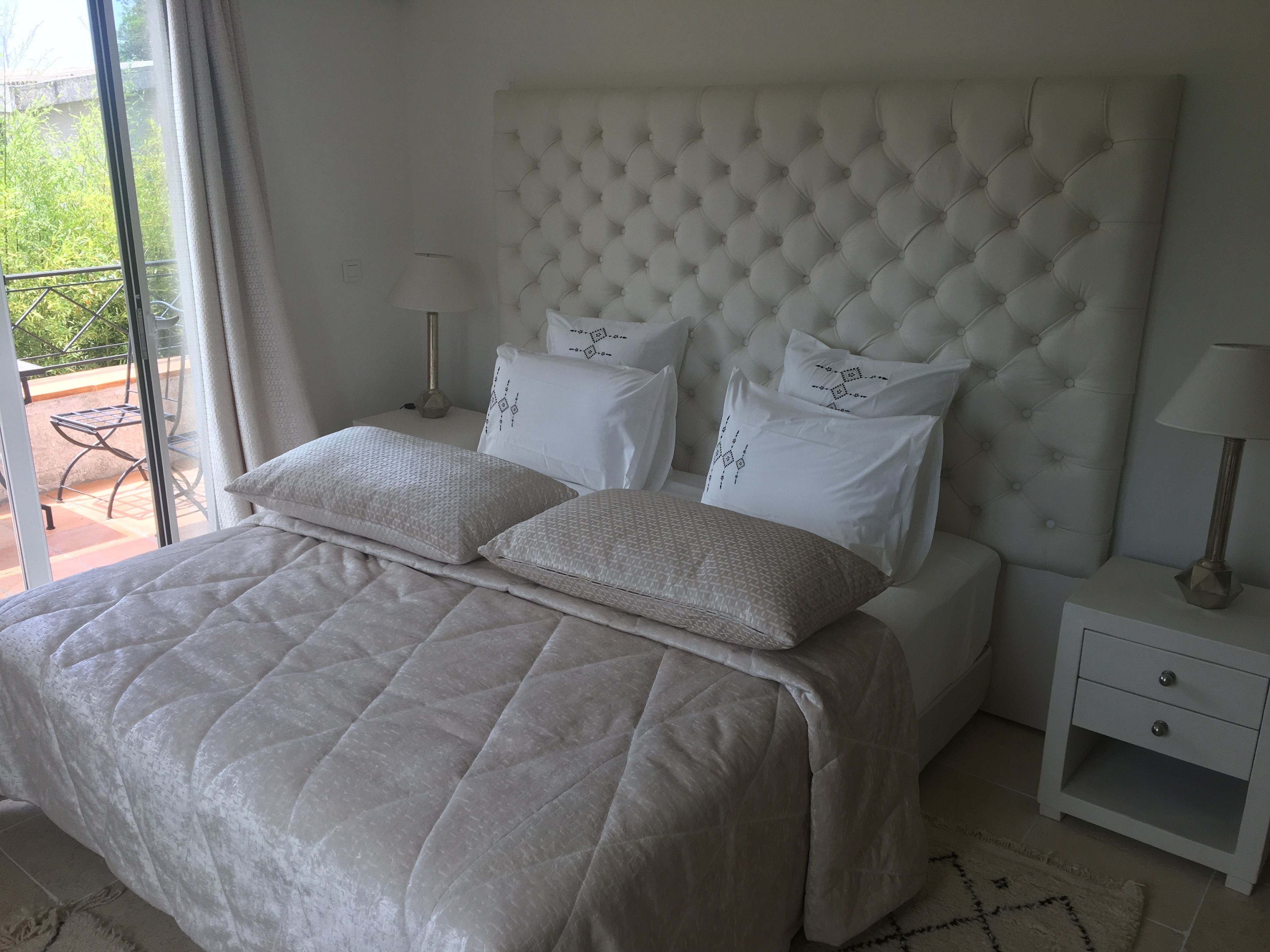 A bedroom with a tufted headboard, a bed with a quilted bedspread, two bedside tables with lamps, and a view of a balcony.