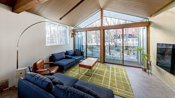 A modern living room with a large sofa, a coffee table, and a floor lamp. There is a large window providing a view of the outdoors. - Grey Wolf Chalet (Hakuba)