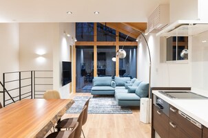Uma cozinha moderna com forno embutido, área de jantar com mesa e cadeiras de madeira, e um sofá azul na sala de estar.