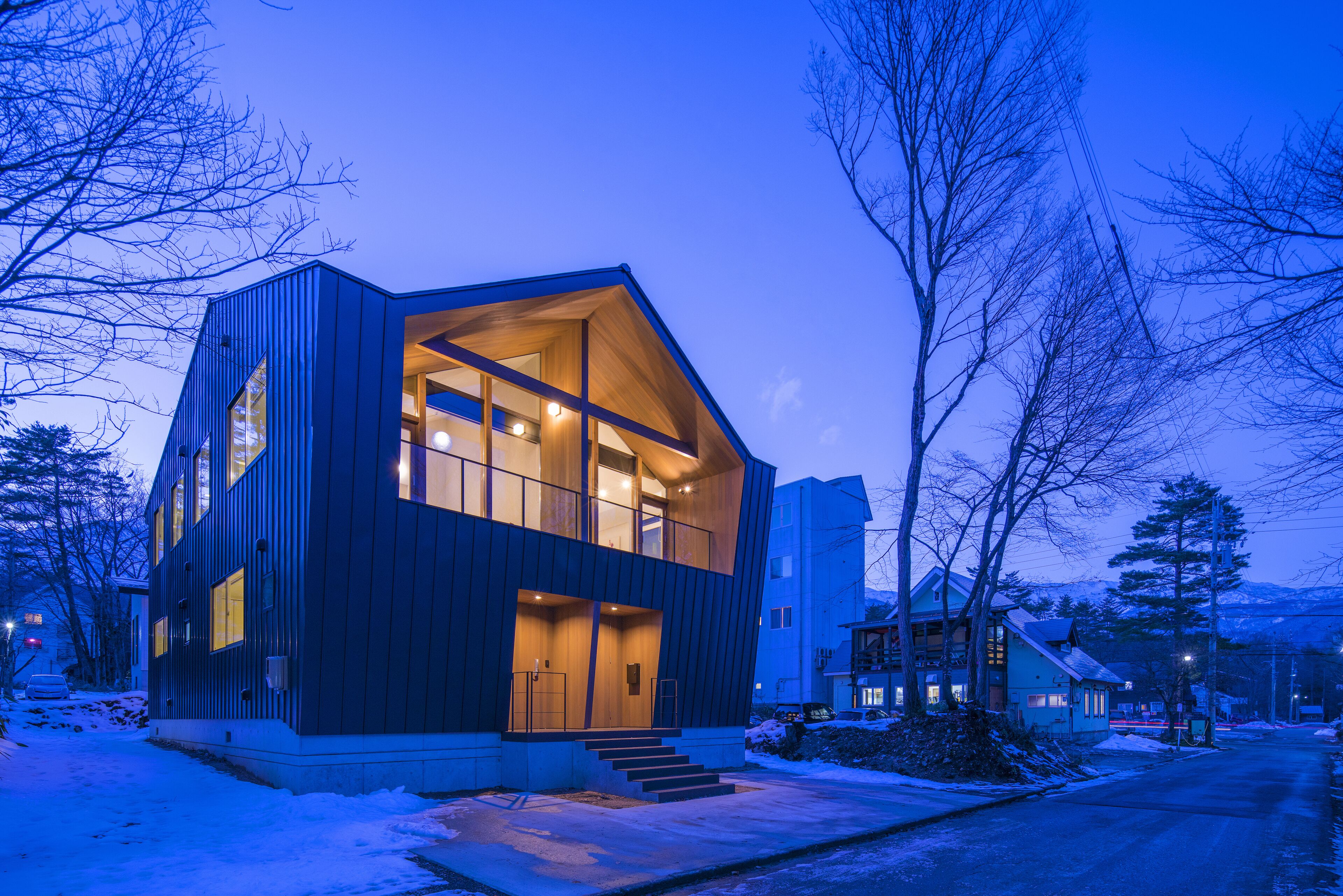 A modern two-story house with a large glass window on the second floor, a wooden exterior, and a snow-covered ground.