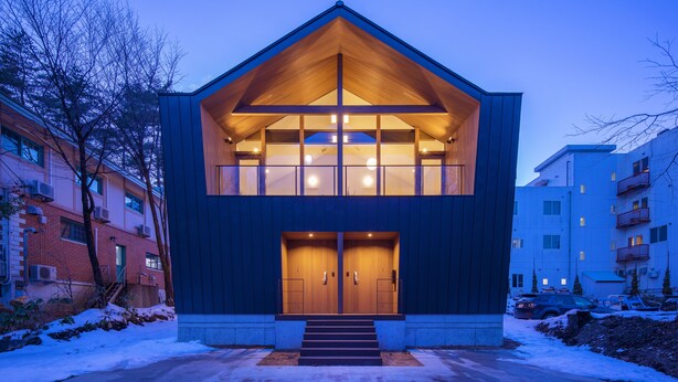 A modern two-story building with a prominent wooden gable roof, large windows, and a set of stairs leading to the entrance. - White Fox Chalet (Hakuba)