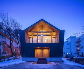 A modern two-story building with a prominent wooden gable roof, large windows, and a set of stairs leading to the entrance. - White Fox Chalet (Hakuba)