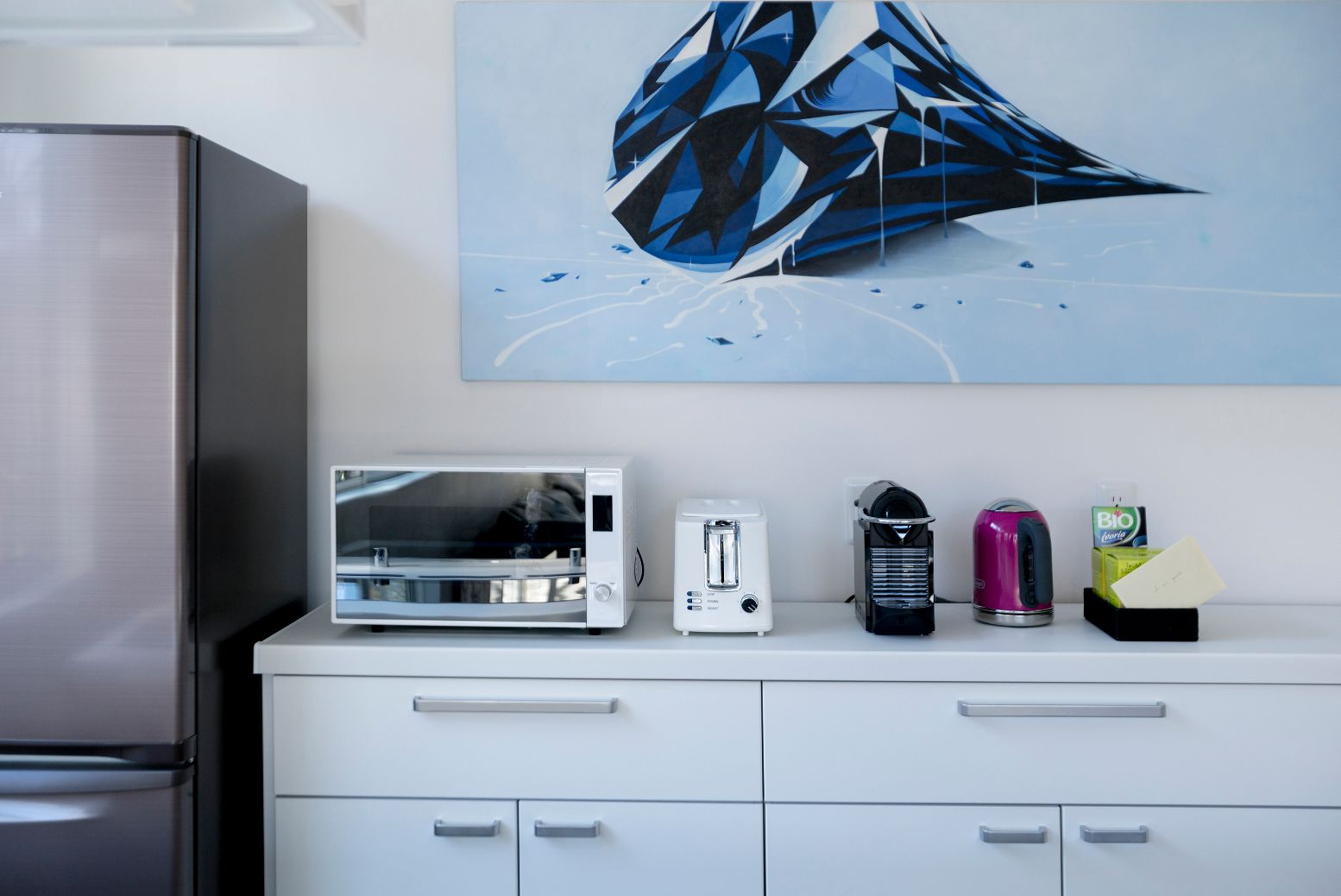 A kitchen with a stainless steel refrigerator, a microwave, a toaster, a coffee maker, and a tea kettle on a white countertop.