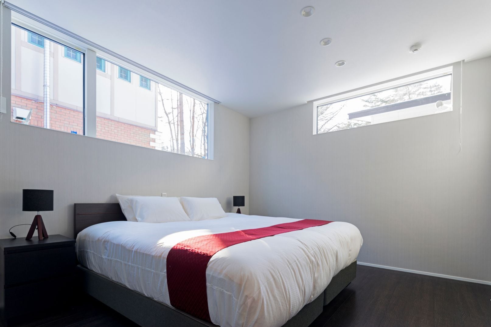 A modern bedroom with a large bed, two bedside tables, and two windows providing natural light.