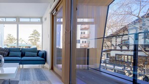 A modern living room with a blue sofa, a glass partition, and a view of a snowy landscape. - White Fox Chalet (Hakuba)