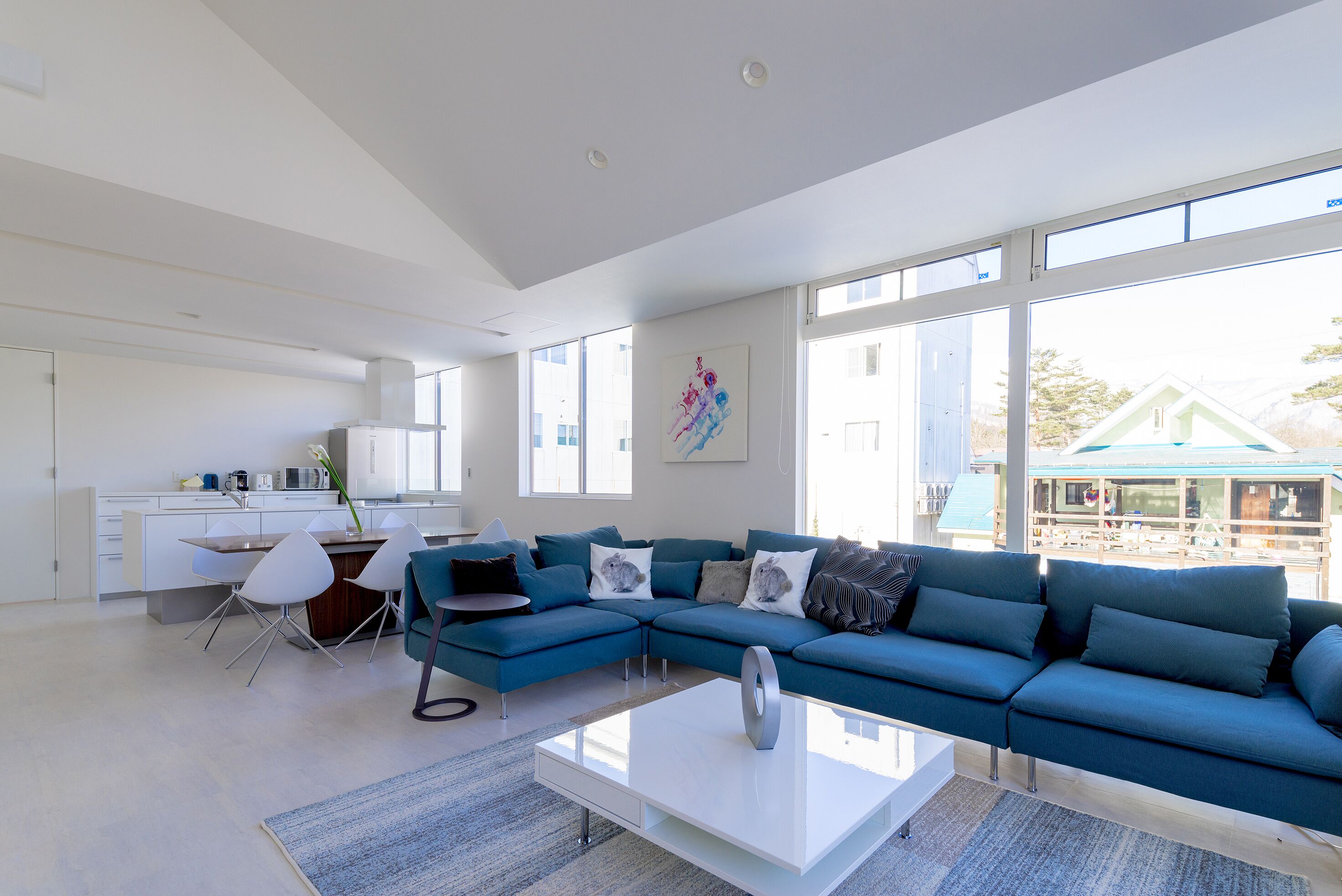 A modern living room with a sectional sofa, a dining area with a table and chairs, and large windows offering a view of the outside.
