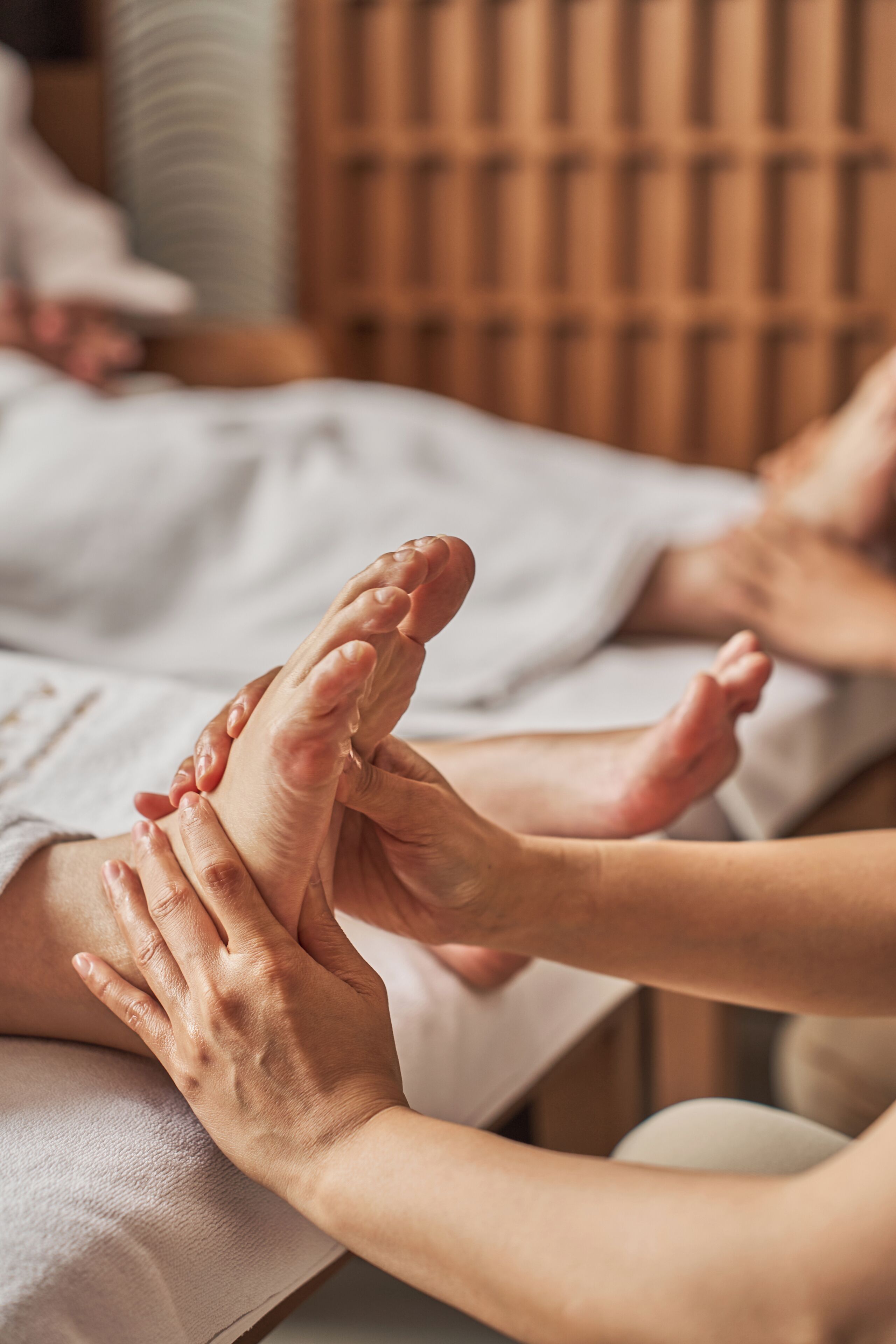 Une séance de massage des pieds dans une pièce dont un mur est recouvert de lambris en bois et où se trouve un lit recouvert d’une courtepointe blanche.