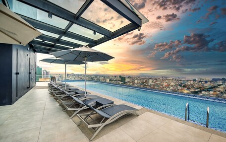 A rooftop pool area with lounge chairs and umbrellas, overlooking a cityscape at sunset.