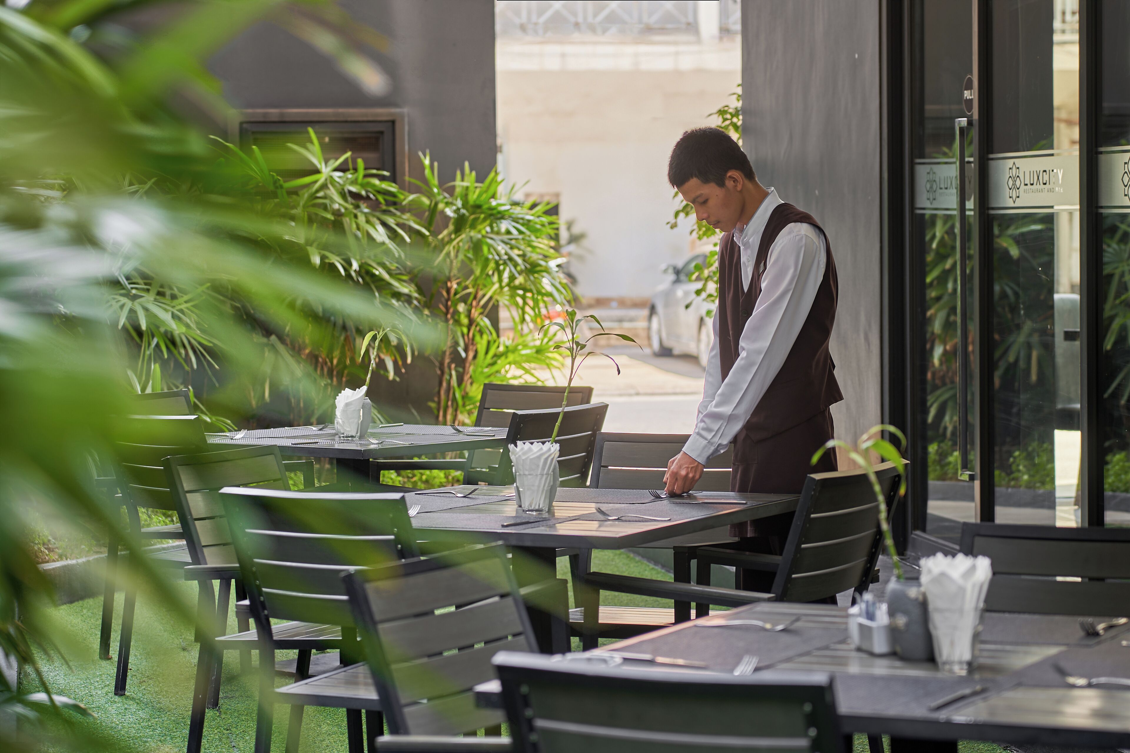 Un espacio de comedor al aire libre con mesas y sillas, un camarero atendiendo una mesa, y un edificio con un letrero que dice "LUXITY".