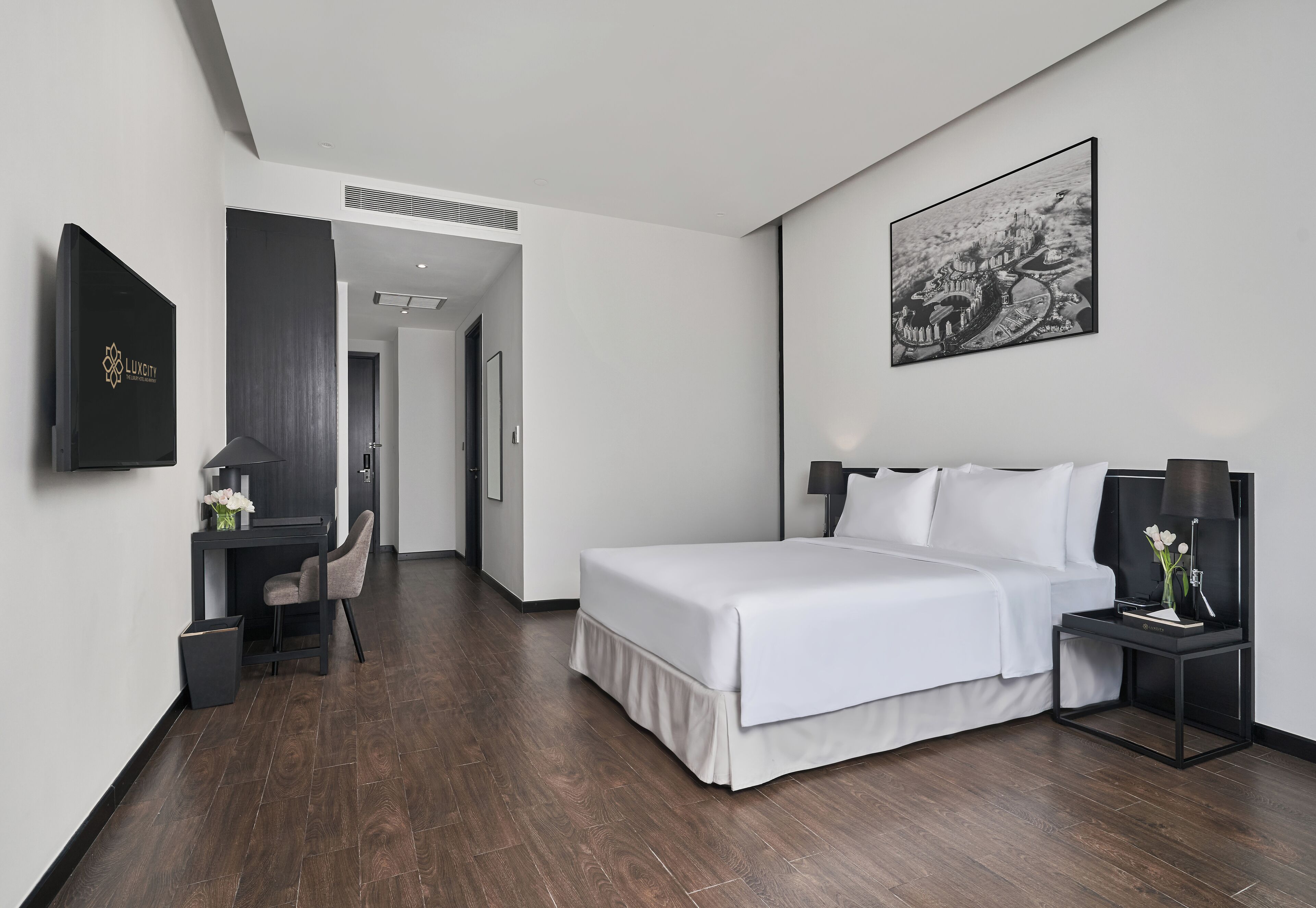 A modern hotel room with a large bed, a desk, and a chair. There is a framed picture on the wall and a TV mounted on the wall.