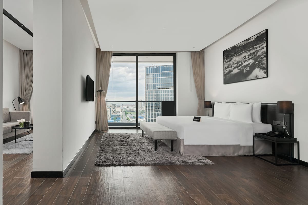 A modern hotel room with a large window offering a city view, a bed with a bedside table, and a flat-screen TV mounted on the wall.
