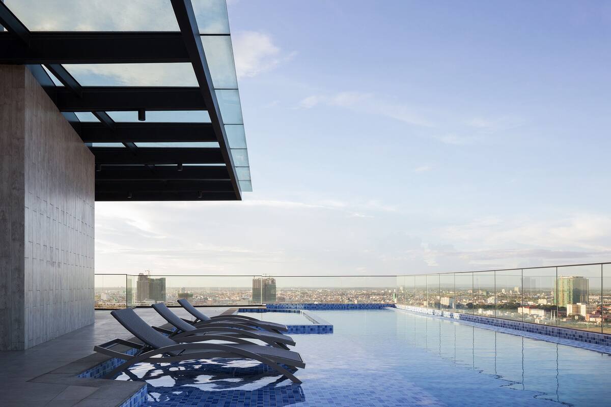 A rooftop infinity pool with lounge chairs, a glass balustrade, and a cityscape view.