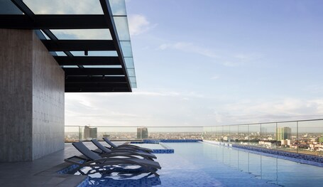 A rooftop infinity pool with lounge chairs, a glass balustrade, and a cityscape view.
