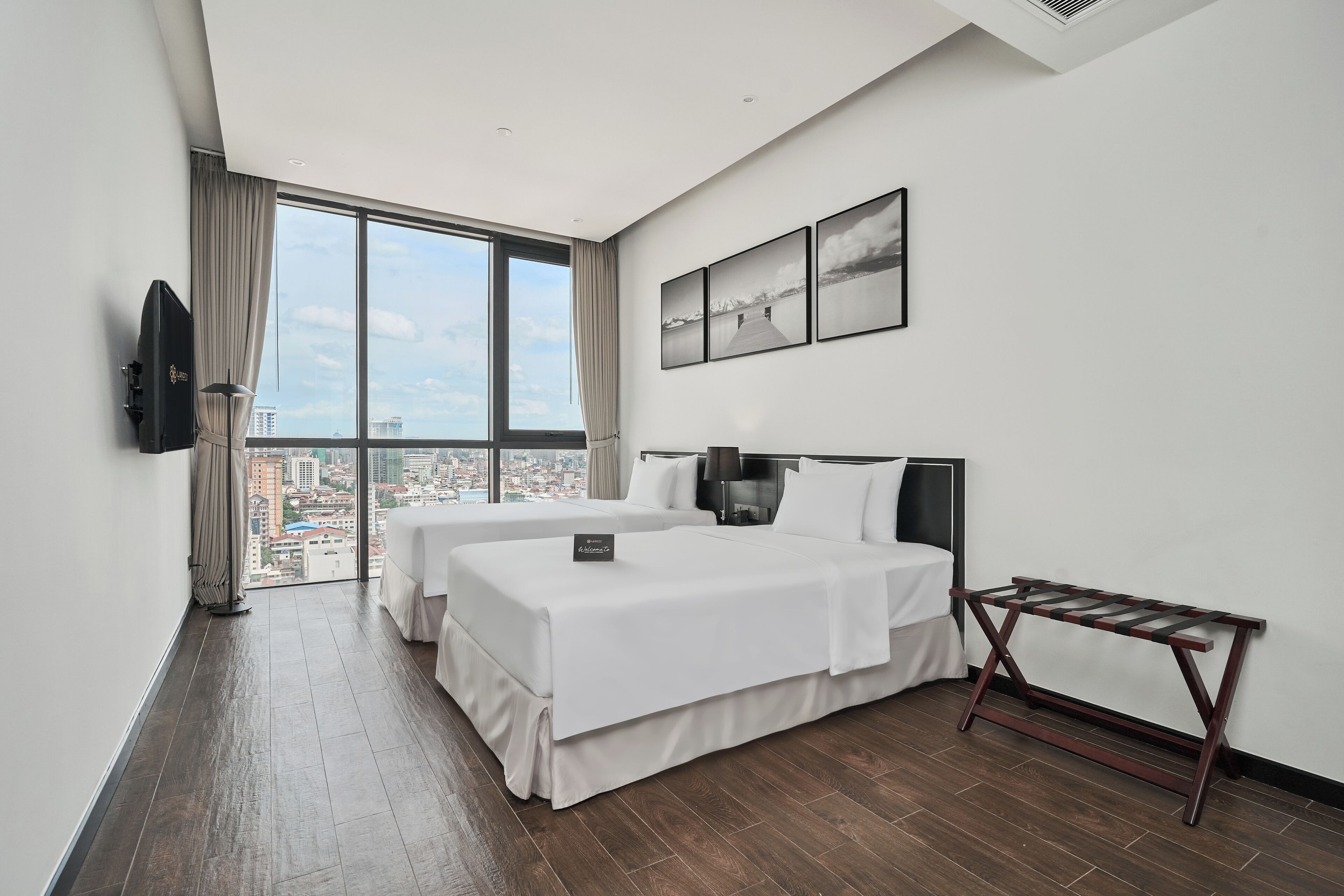 A hotel room with two beds, a large window with a city view, a flat-screen TV, and a small table.