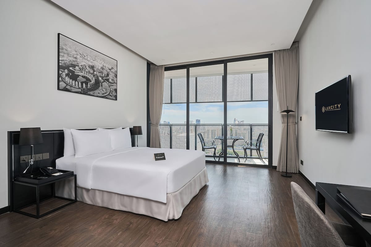 A modern hotel room with a large bed, a desk with a lamp, a TV, and a balcony with a city view.