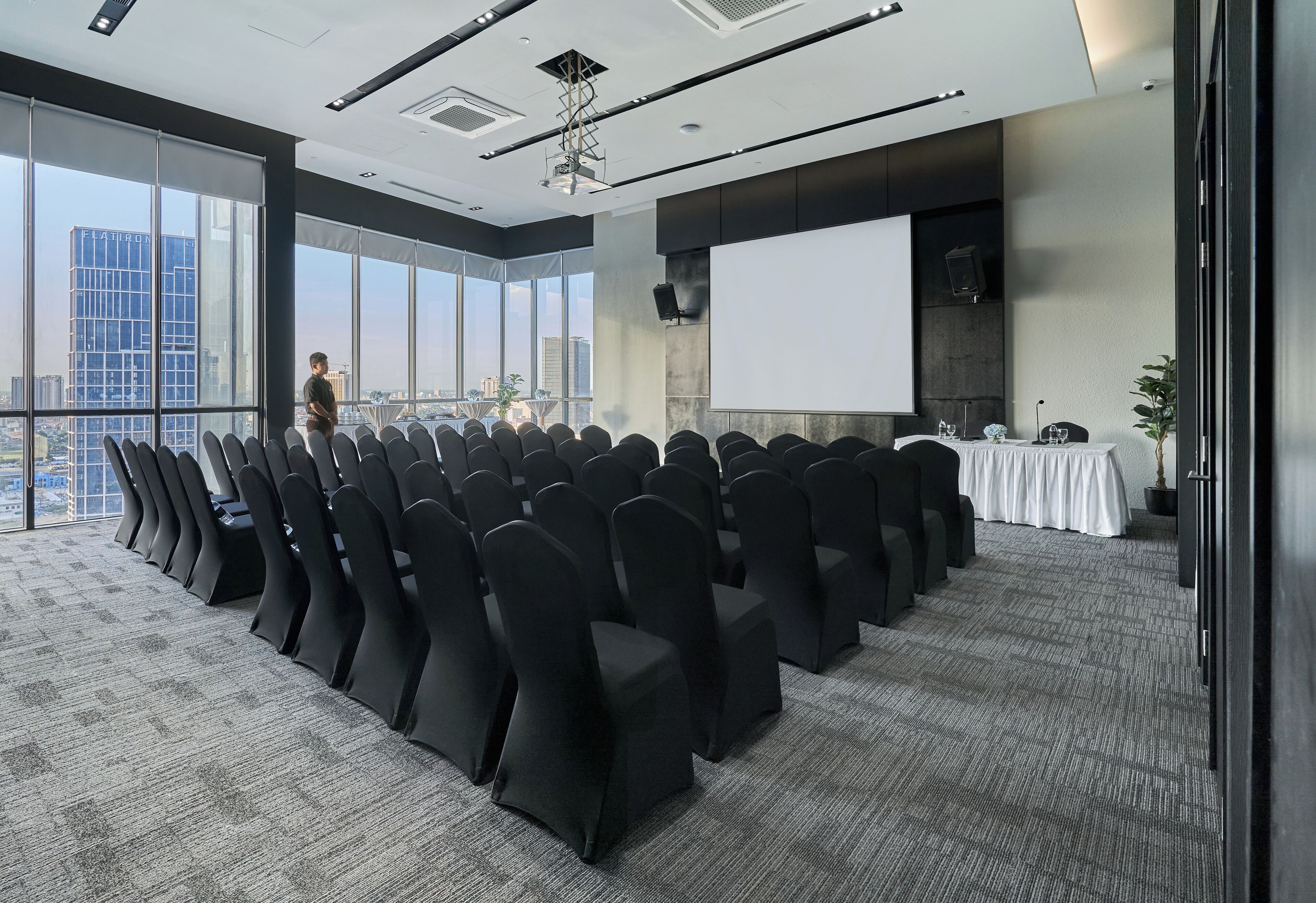 A conference room with rows of chairs, a projector screen, and a large window with a city view.