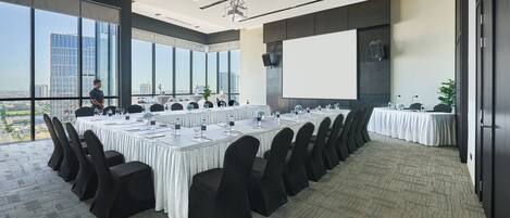 Un salón de conferencias con una gran mesa dispuesta para una reunión, amplias ventanas que dan a una vista de la ciudad y una pantalla de proyector.