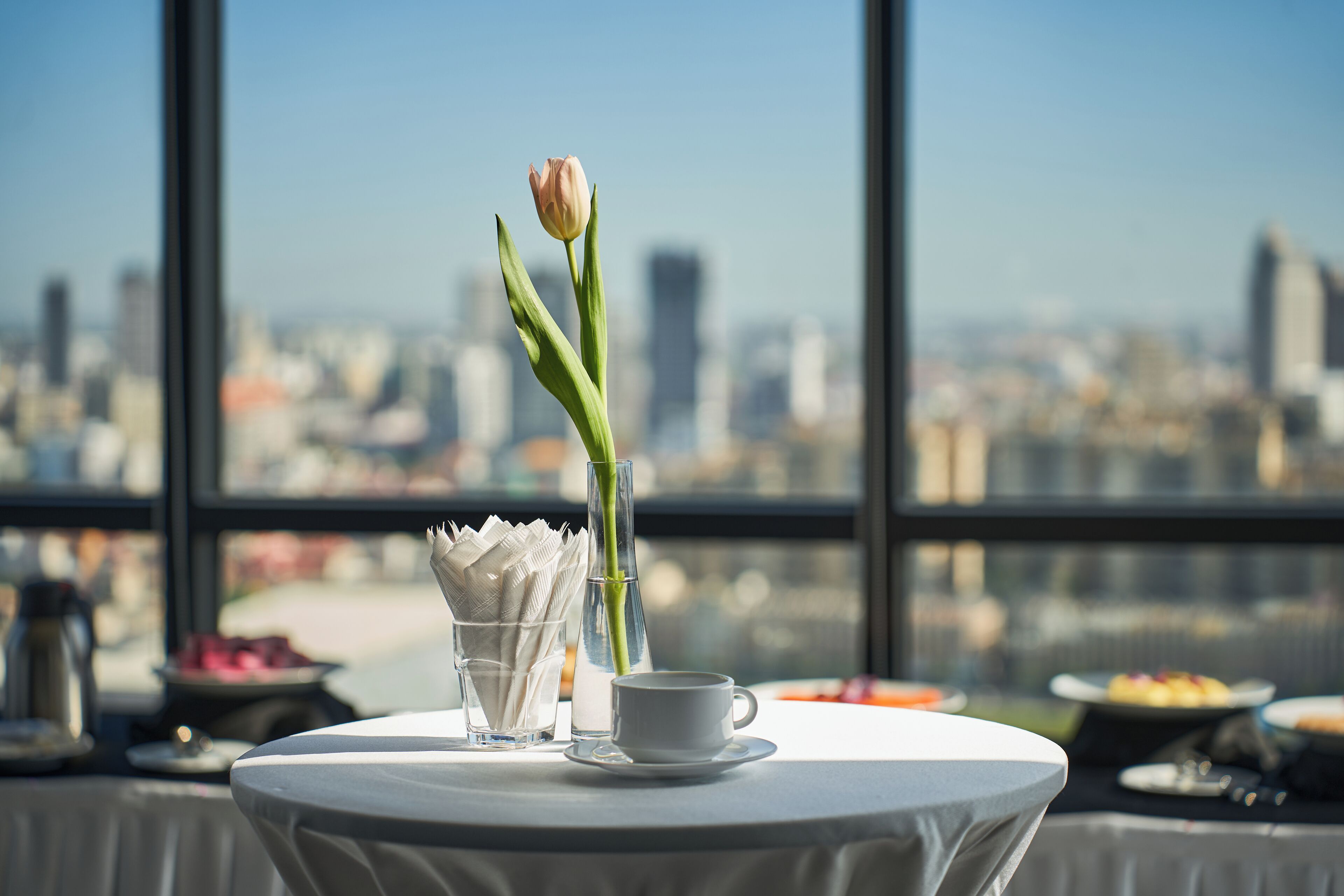 Una mesa con una taza, un florero con una sola flor y un vaso de agua, con vista a un paisaje urbano.
