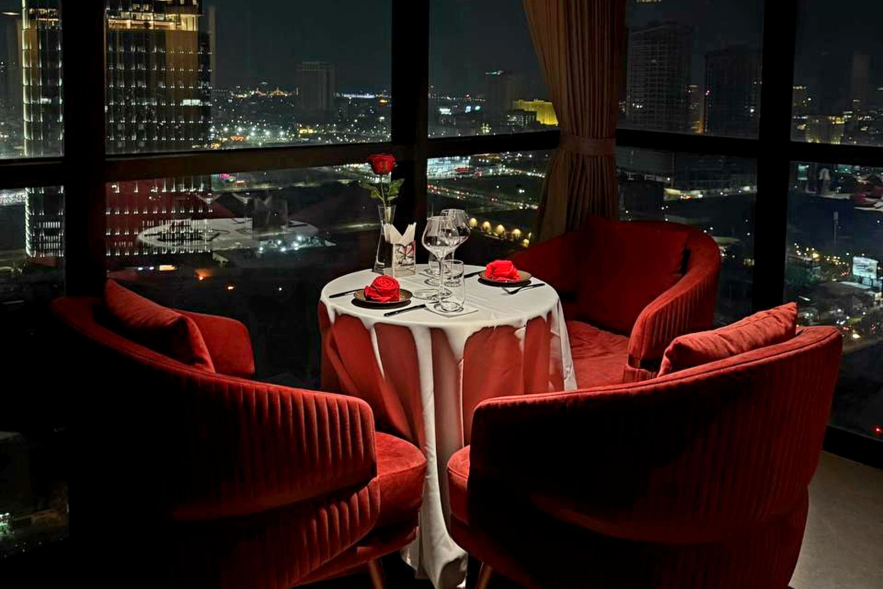 A dining table with four red chairs, a white tablecloth, and a vase with a single red rose. The table is set with a wine glass, a water glass, and a red napkin.