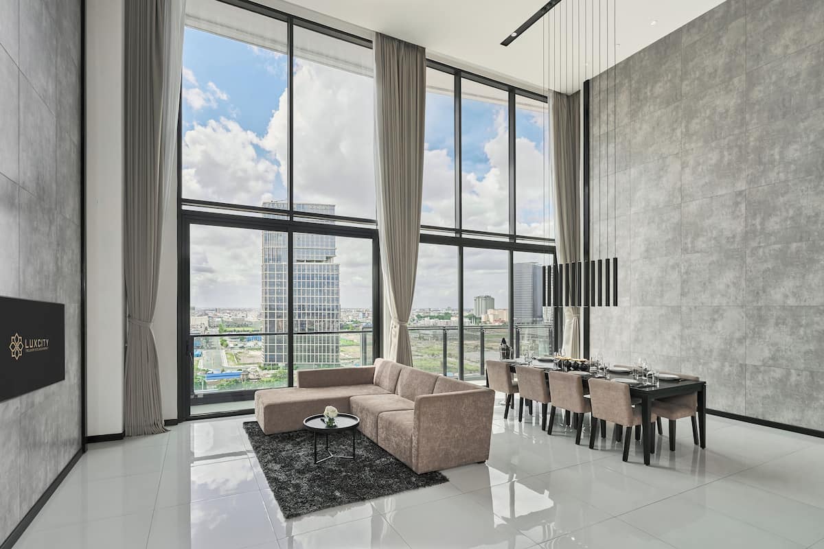 A modern living room with a large window, a sofa, a dining table set, and a flat-screen TV mounted on the wall.