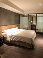 Una habitación de hotel moderna con una cama grande, una mesita de noche, un escritorio y una zona de ducha con paredes de vidrio.