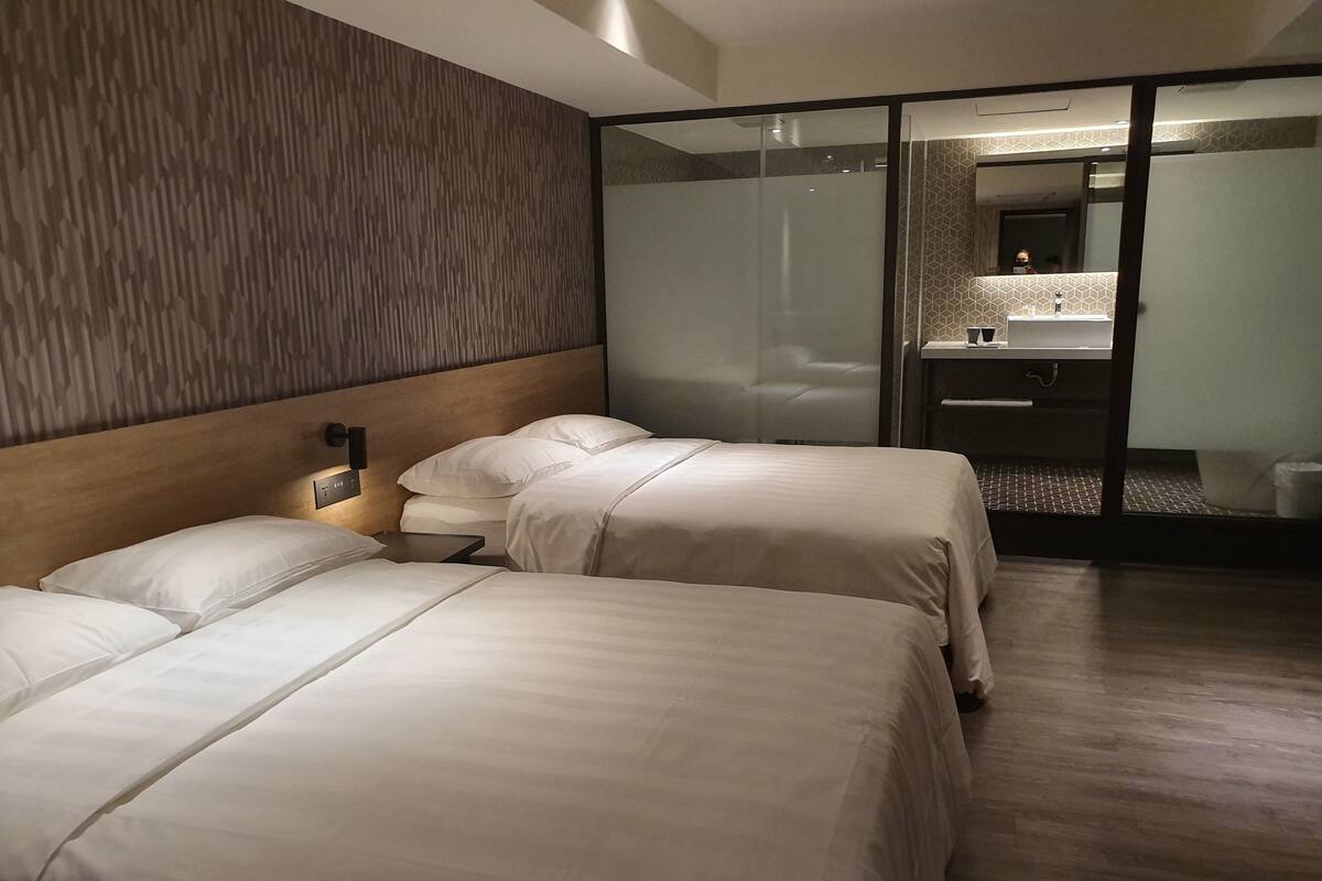 Une chambre d’hôtel avec deux lits, une tête de lit en bois, une cloison en verre et une salle de bain équipée d’un miroir et d’un lavabo.