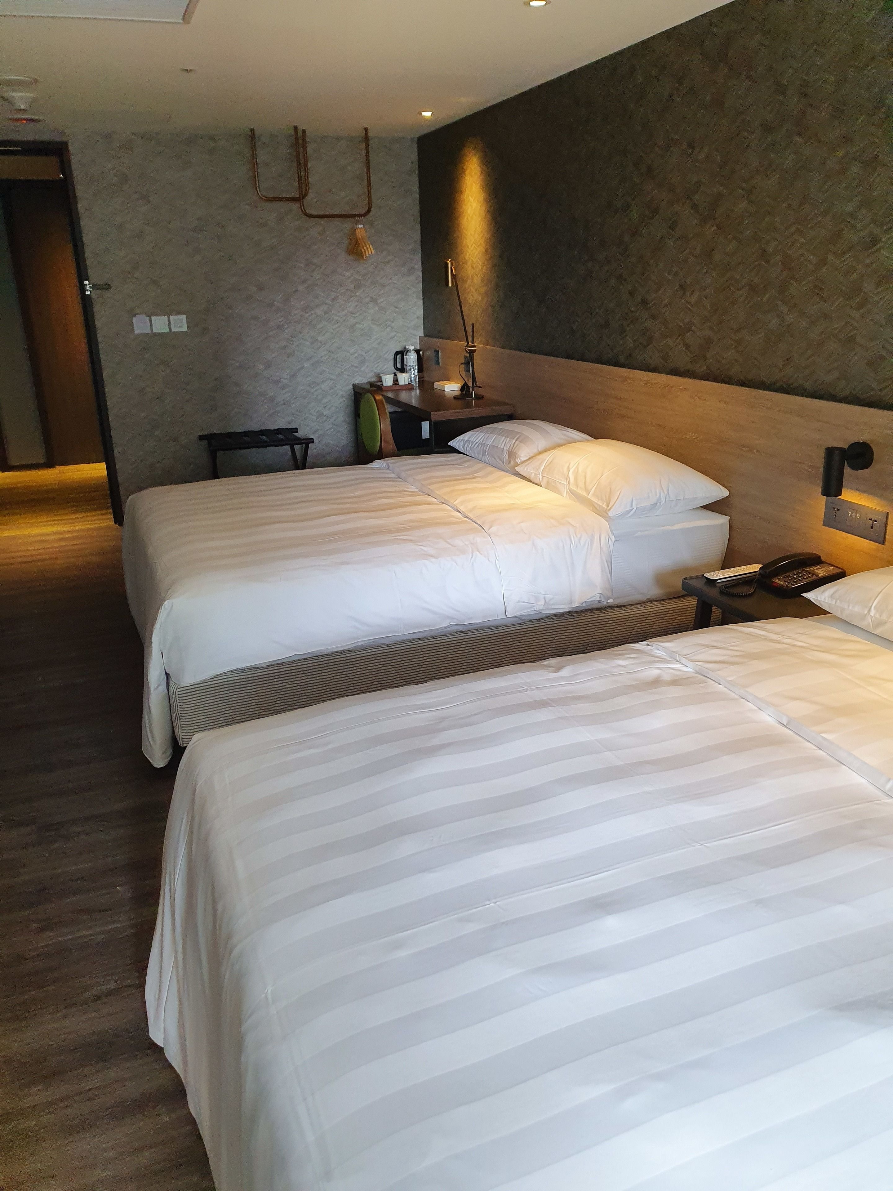 A hotel room with two beds, a bedside table with a lamp, a wall-mounted phone, and a small table with a kettle and cups.