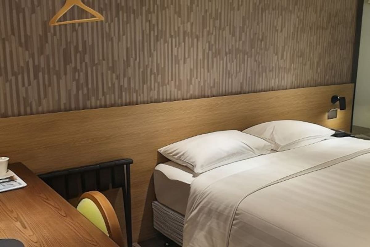 A hotel room with a wooden headboard, a bed with white bedding, a bedside table with a cup and a book, and a chair with a yellow cushion.