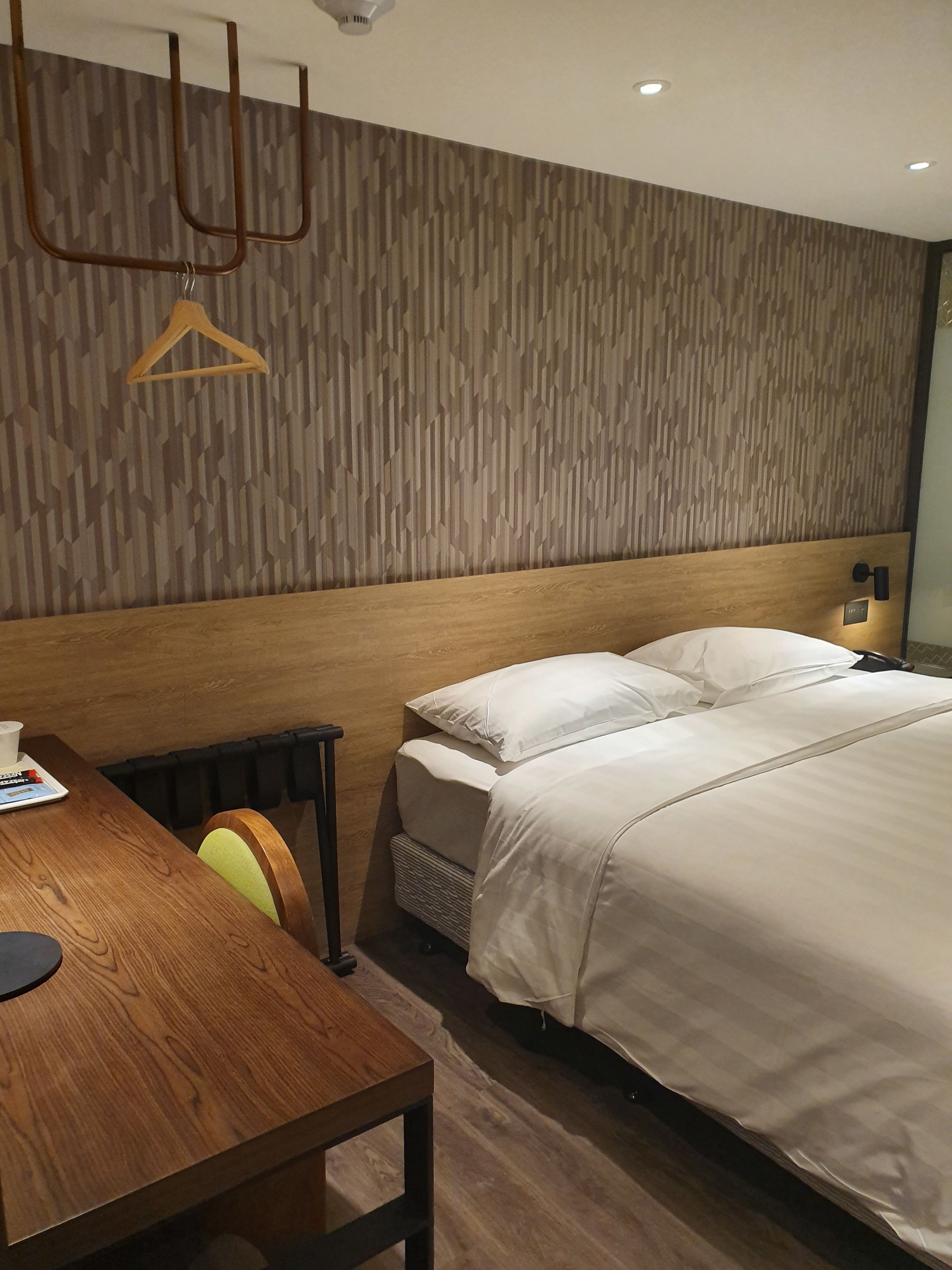 A modern hotel room with a wooden headboard, a bed with white bedding, a wooden table with a chair, and a hanging light fixture.
