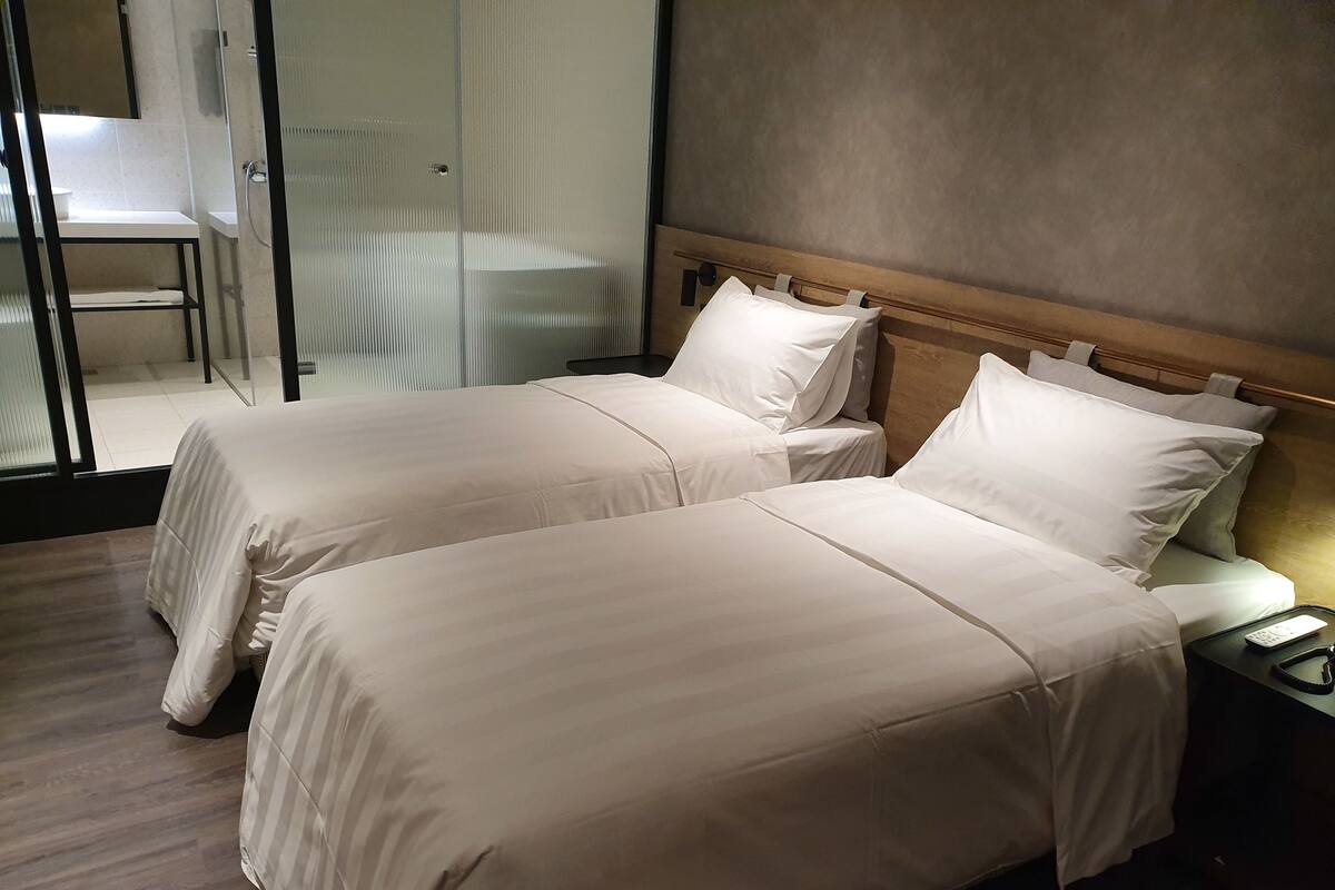 Une chambre d’hôtel avec deux lits, une table de chevet avec une télécommande et une cloison en verre séparant la chambre d’un espace salle de bain.