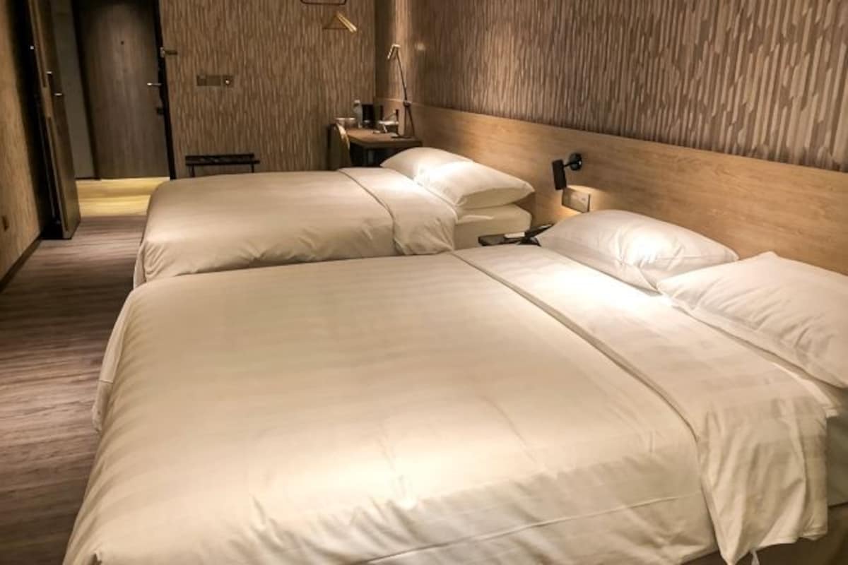 A hotel room with two beds, a wooden headboard, a bedside table with a lamp, and a small seating area.