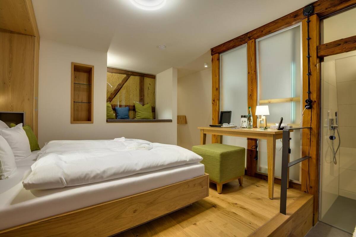 A modern bedroom with a wooden bed, a small desk, a green chair, and a shower area.
