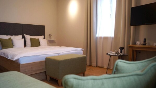 Habitación de hotel con cama, una mesita con teléfono y cafetera, televisión montada en la pared y una ventana con cortinas.