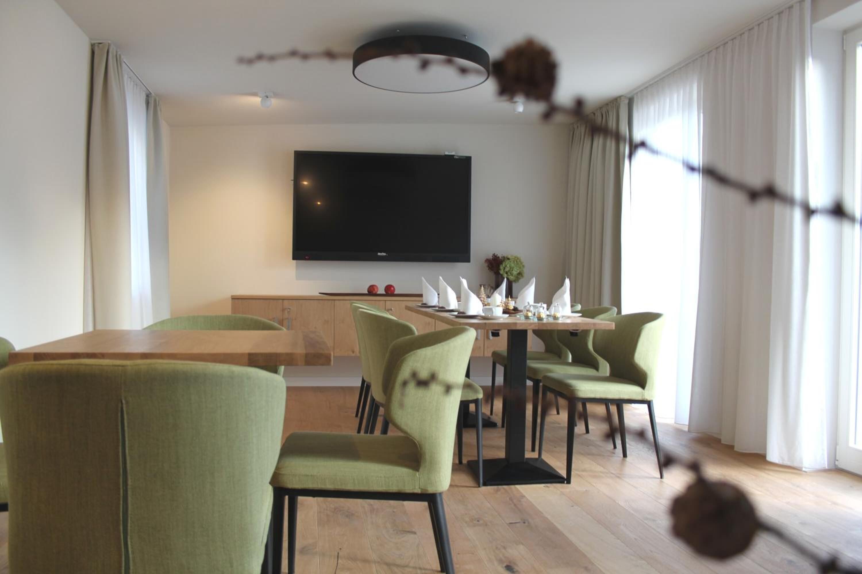 Uma sala de jantar moderna com mesa de madeira e cadeiras estofadas em verde, uma TV de tela plana na parede e uma janela com cortinas.