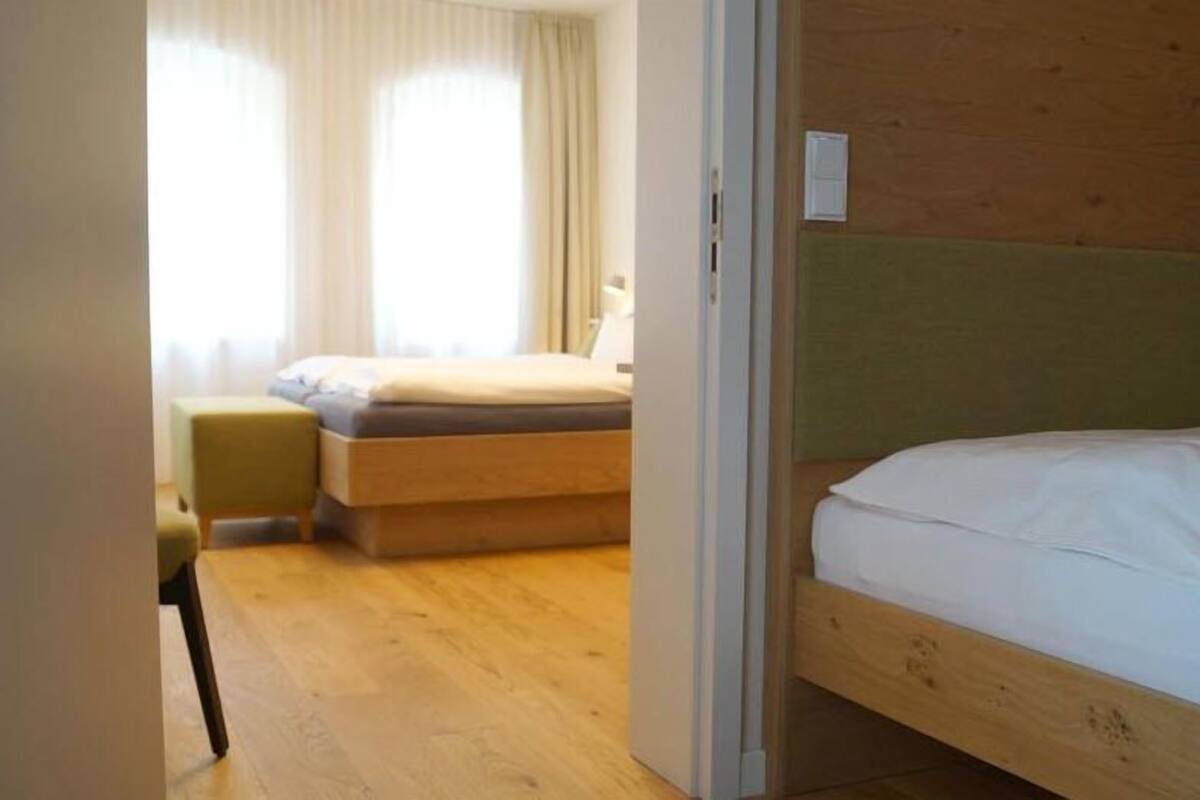A hotel room with a wooden headboard bed, a smaller bed with a green chair, and a window with curtains.