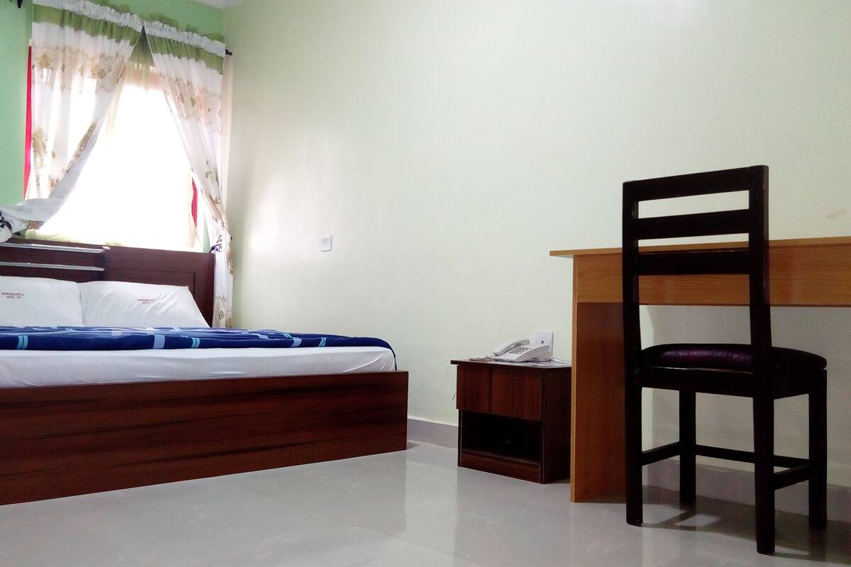 A bedroom with a wooden bed, a desk with a chair, a telephone, and a window with curtains.