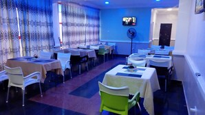 A restaurant with multiple tables and chairs, a television mounted on the wall, and a fan on the floor. - Denposon Royal Hotel (Lagos)