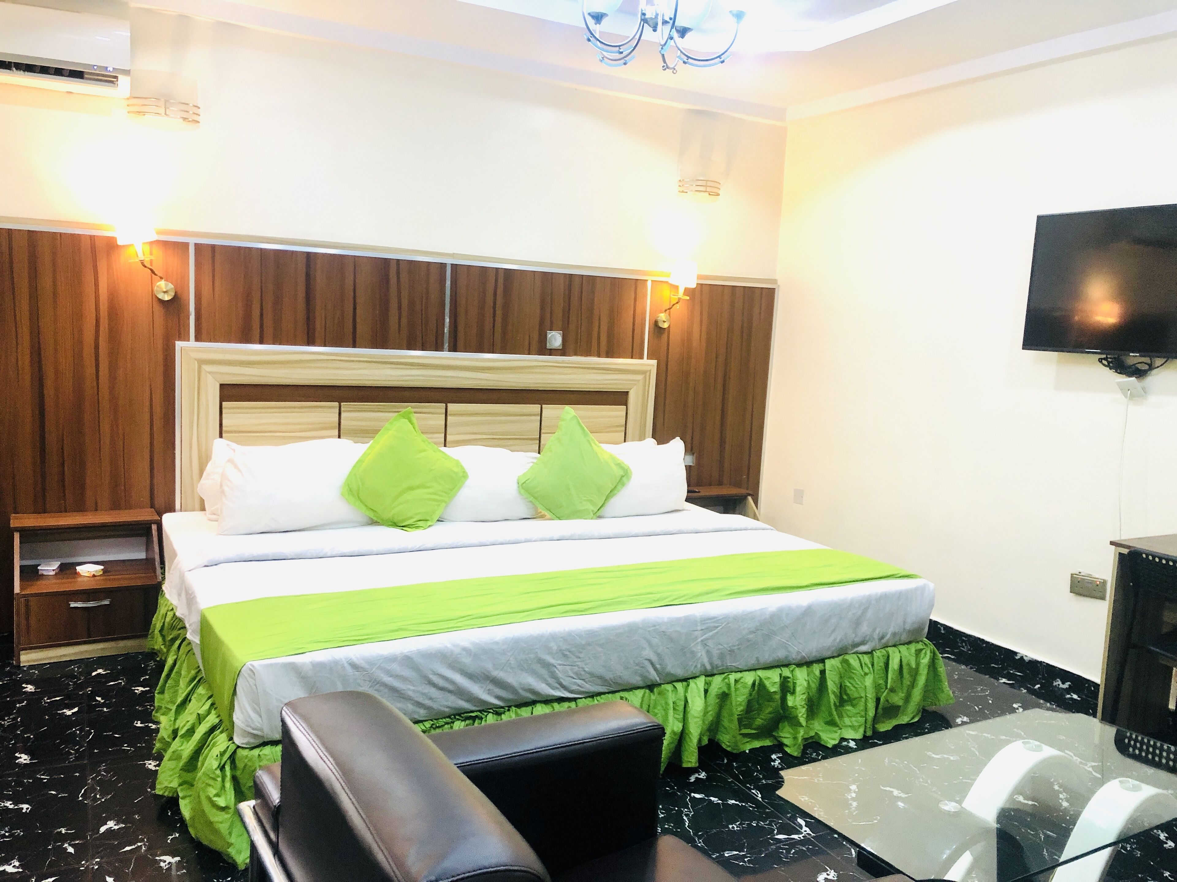 A hotel room with a bed, green pillows, a green bedspread, a wooden headboard, a television on the wall, and a small table with a chair.