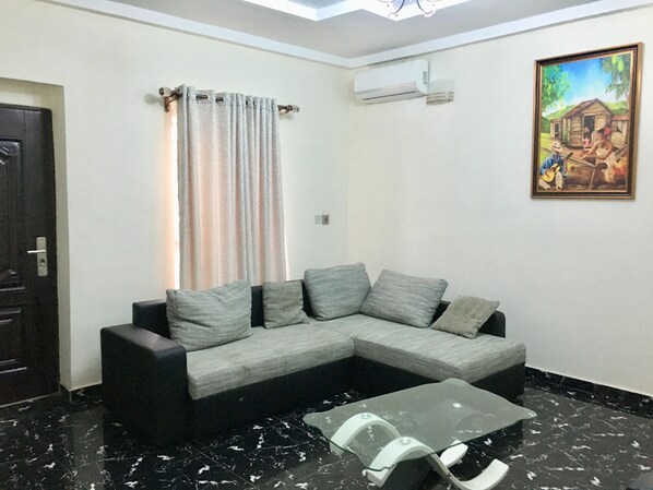 A living room with a grey sectional sofa, a glass coffee table, a dark door, a window with curtains, an air conditioning unit, and a painting on the wall. - Sky Rock Hotels (Lagos)