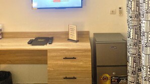 Una habitación de hotel con una televisión mostrando el canal Cartoon Network, un escritorio de madera con un menú, un minibar y un refrigerador Neón.