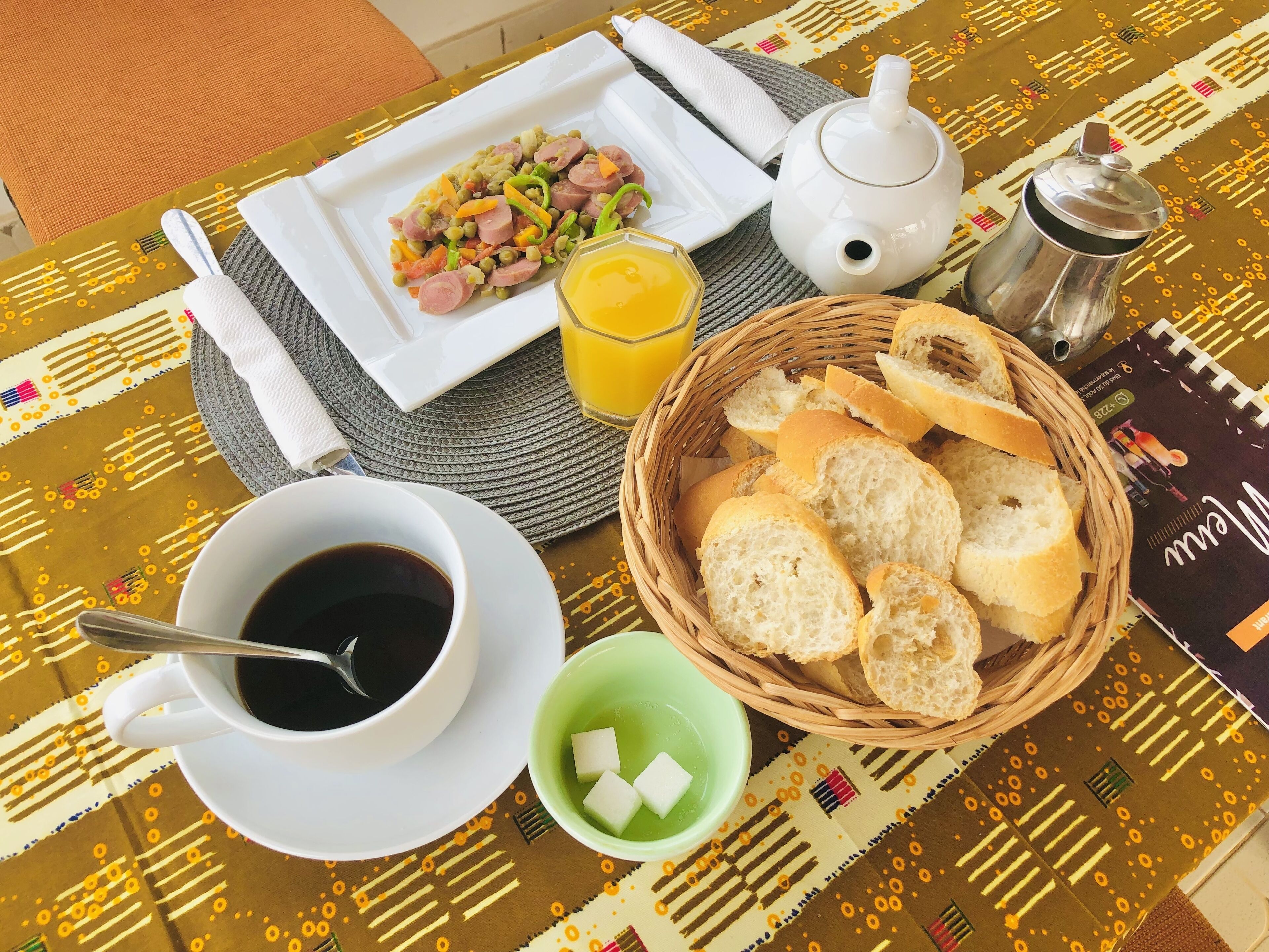 A breakfast setting with a plate of food, a cup of coffee, a glass of orange juice, a teapot, a sugar bowl, and a basket of bread.