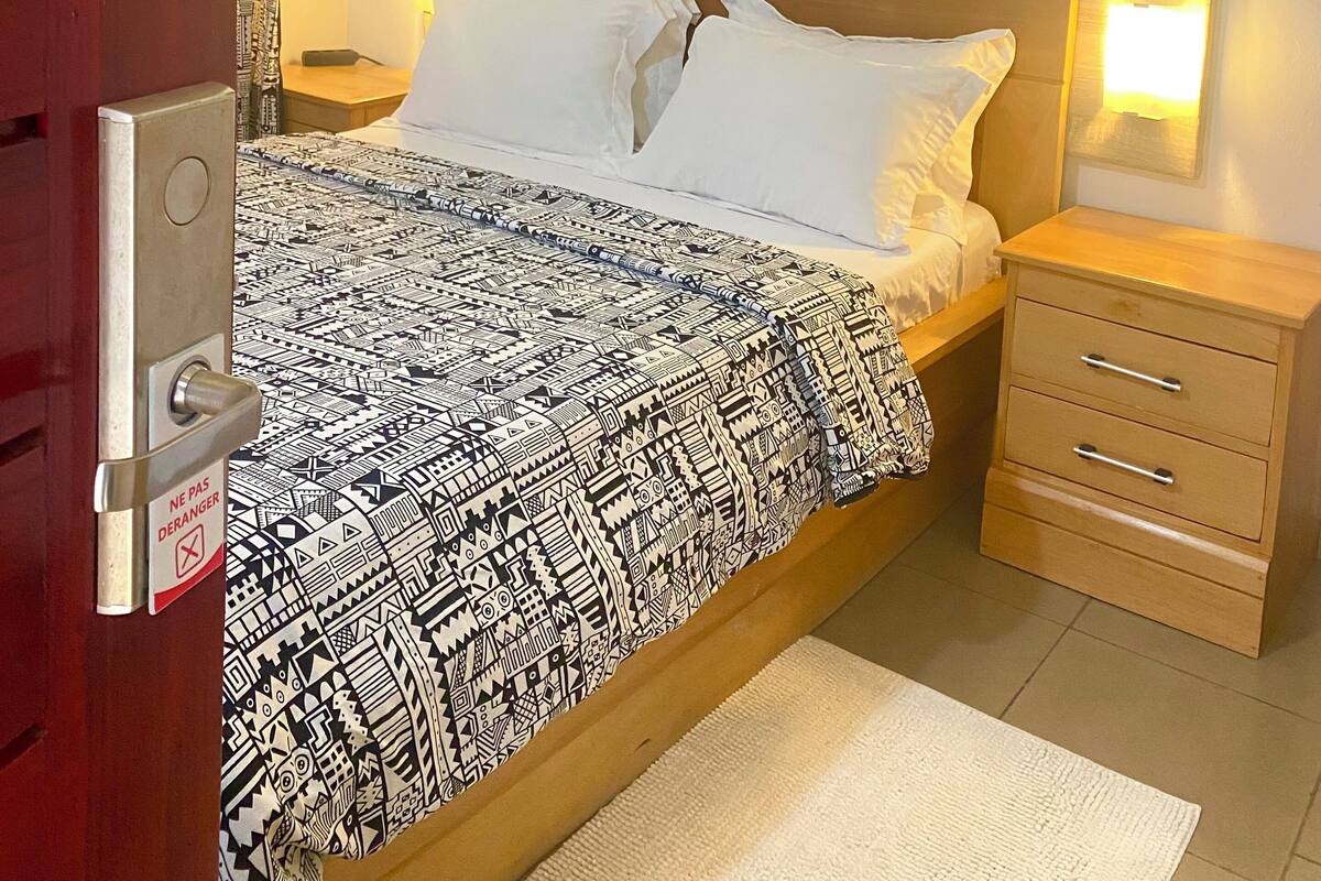 Una cama bien tendida con ropa de cama estampada, una mesita de noche de madera con cajones, un cuadro colgado en la pared y una puerta con cerradura de seguridad.