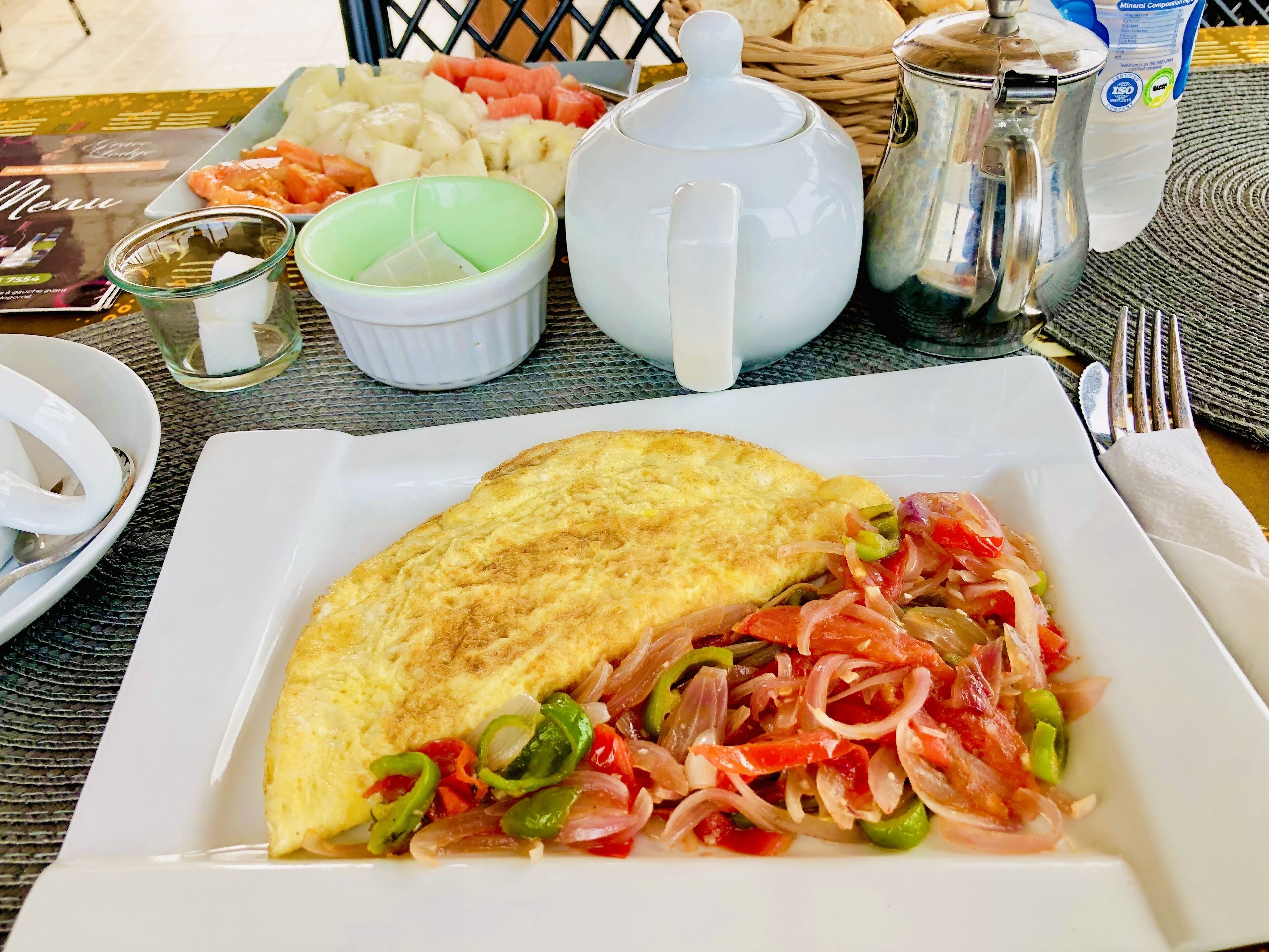 A meal with an omelette and a vegetable salad on a white plate, accompanied by a teapot, a jug, and a basket of bread.