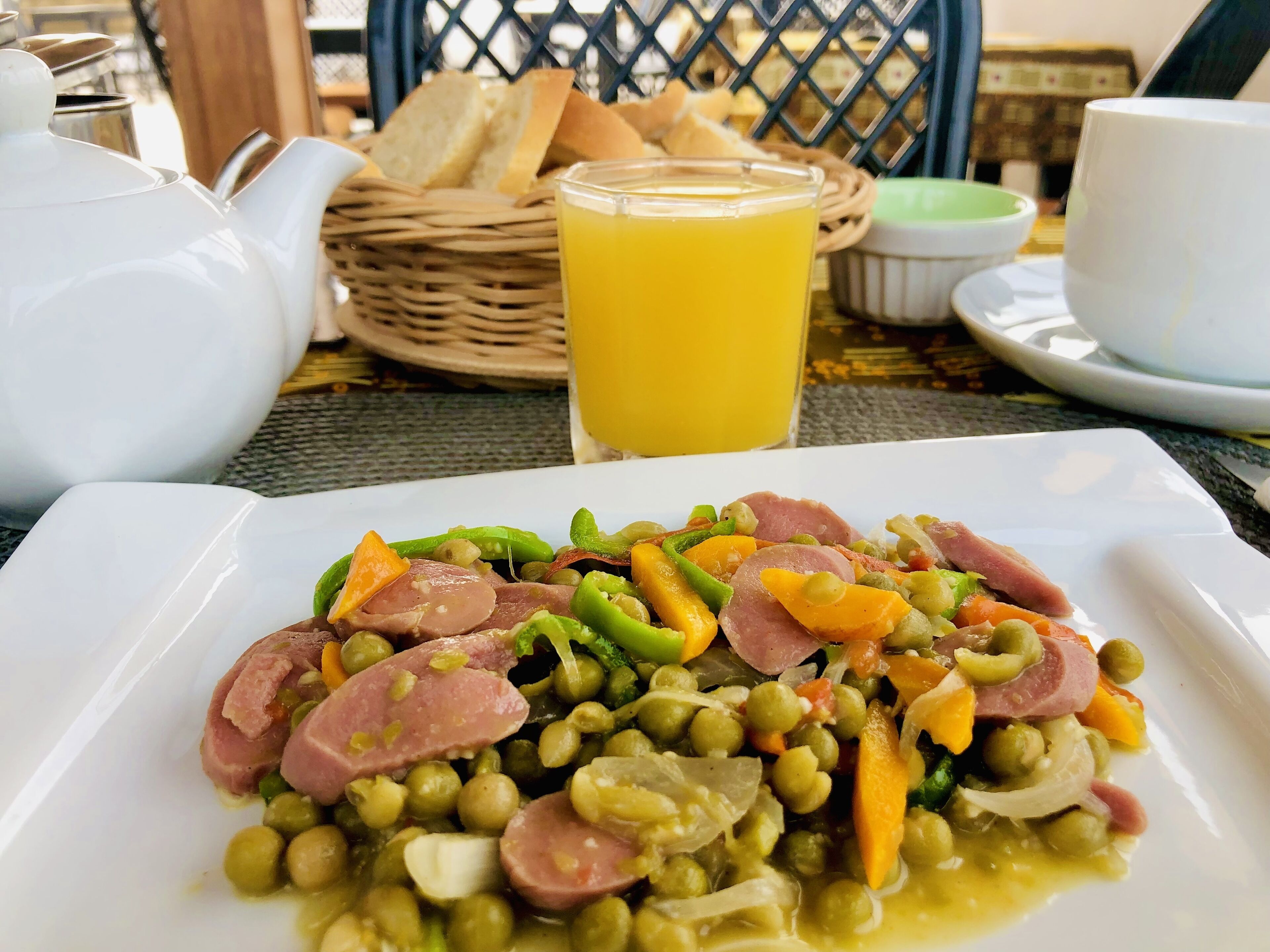A meal with peas, carrots, and sausage on a white plate, accompanied by a glass of orange juice, a teapot, and a basket of bread.