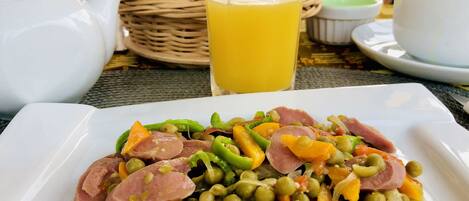 A meal with peas, carrots, and sausage on a white plate, accompanied by a glass of orange juice, a teapot, and a basket of bread.